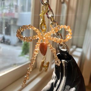 Keychain with decorative bow and 'H' charm attached to a black bag by a window.